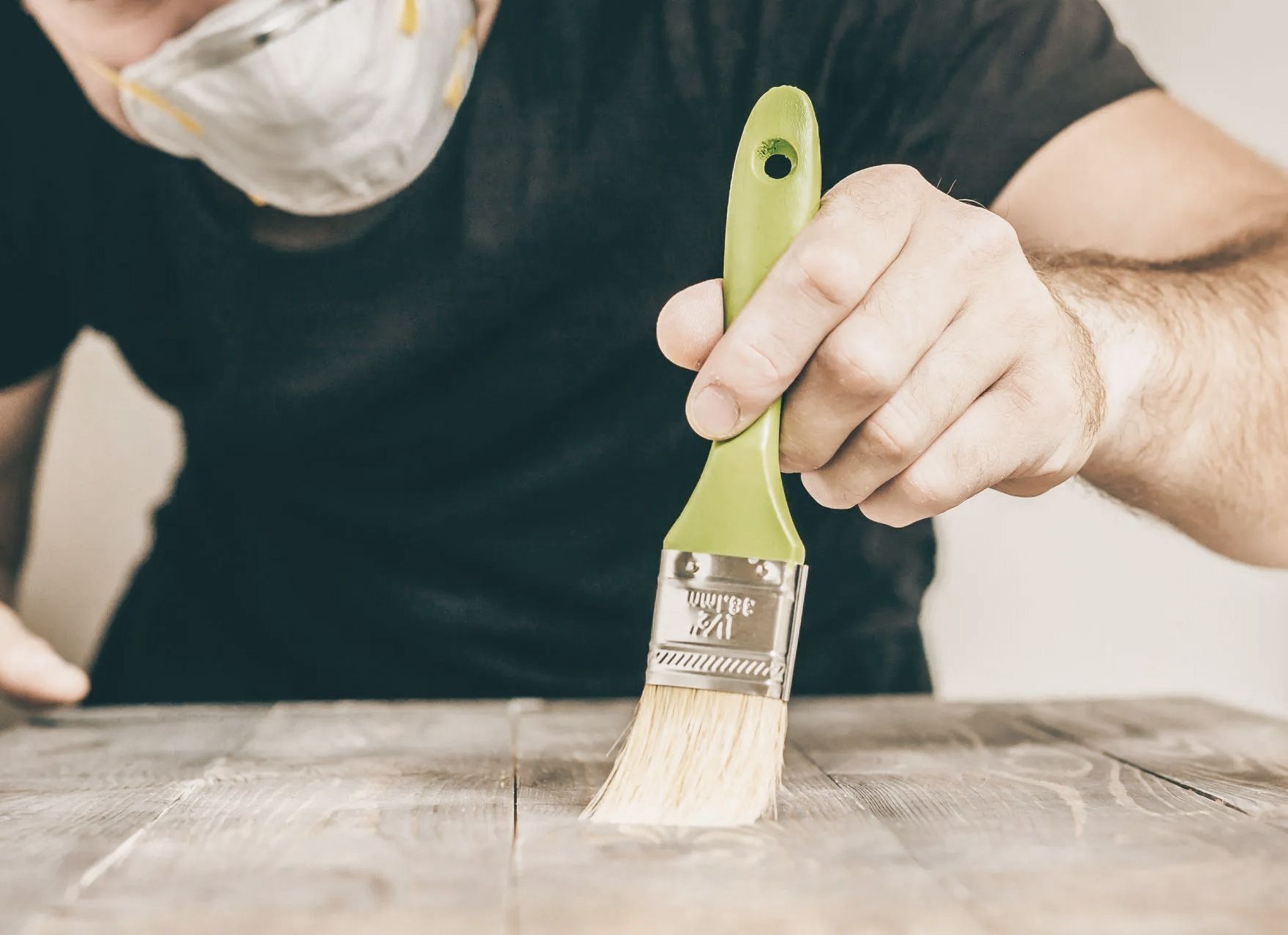 Person wearing mask applying something with a paintbrush to a wooden surface.