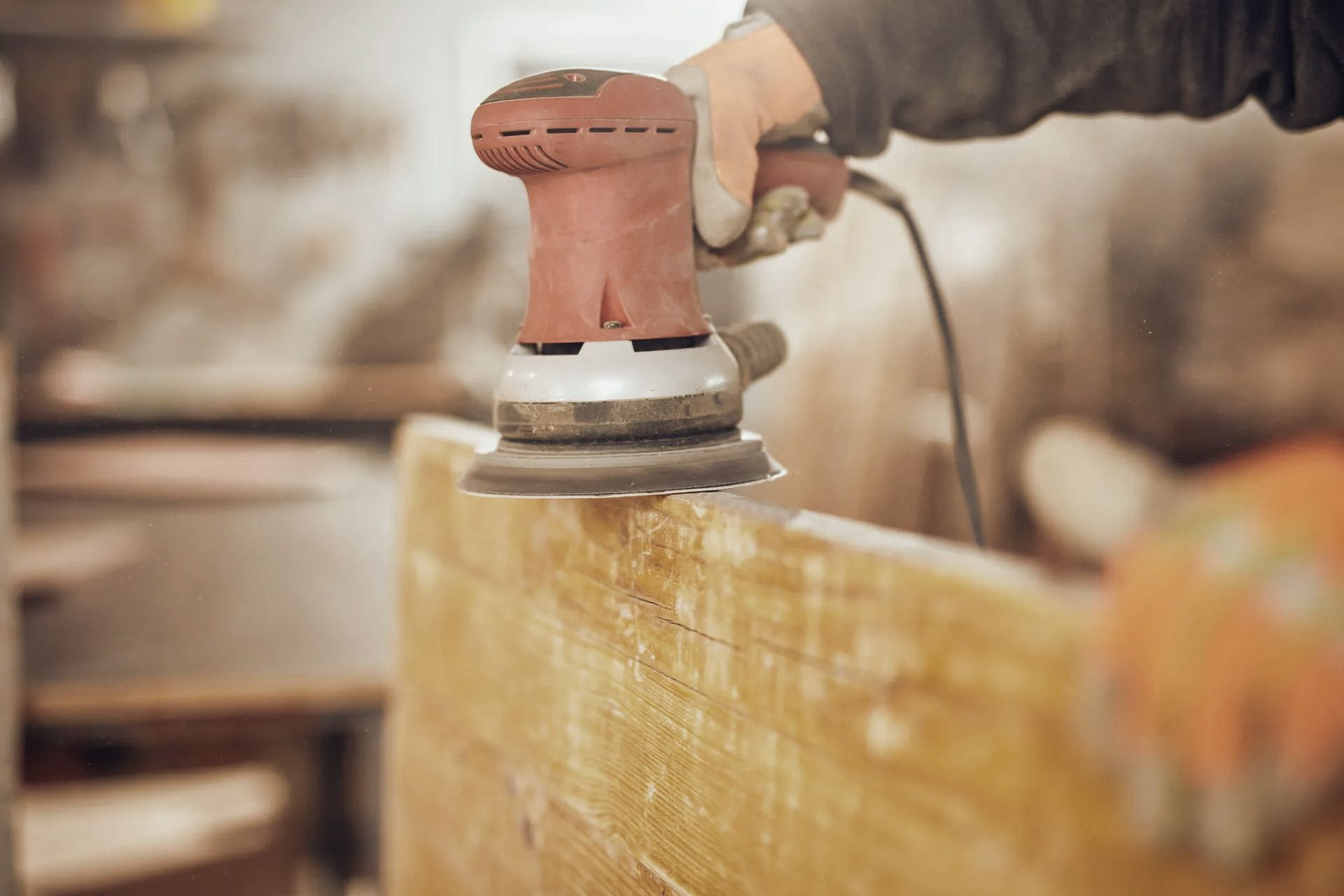 Person sanding a wooden surface with a red electric sander in a workshop.