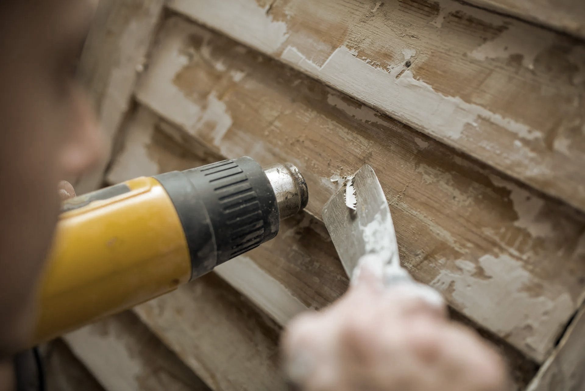 Person using a heat gun and scraper to remove paint from a wooden surface.