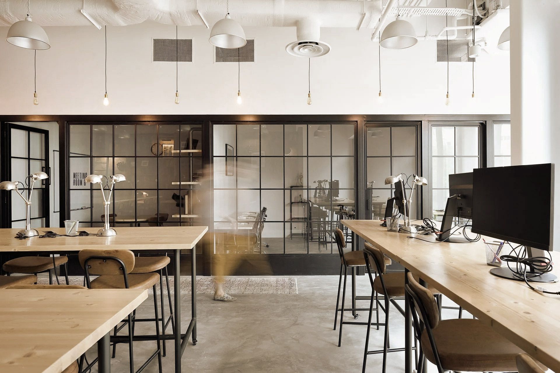 Modern office with long wooden desks, glass-walled rooms, and hanging lights.