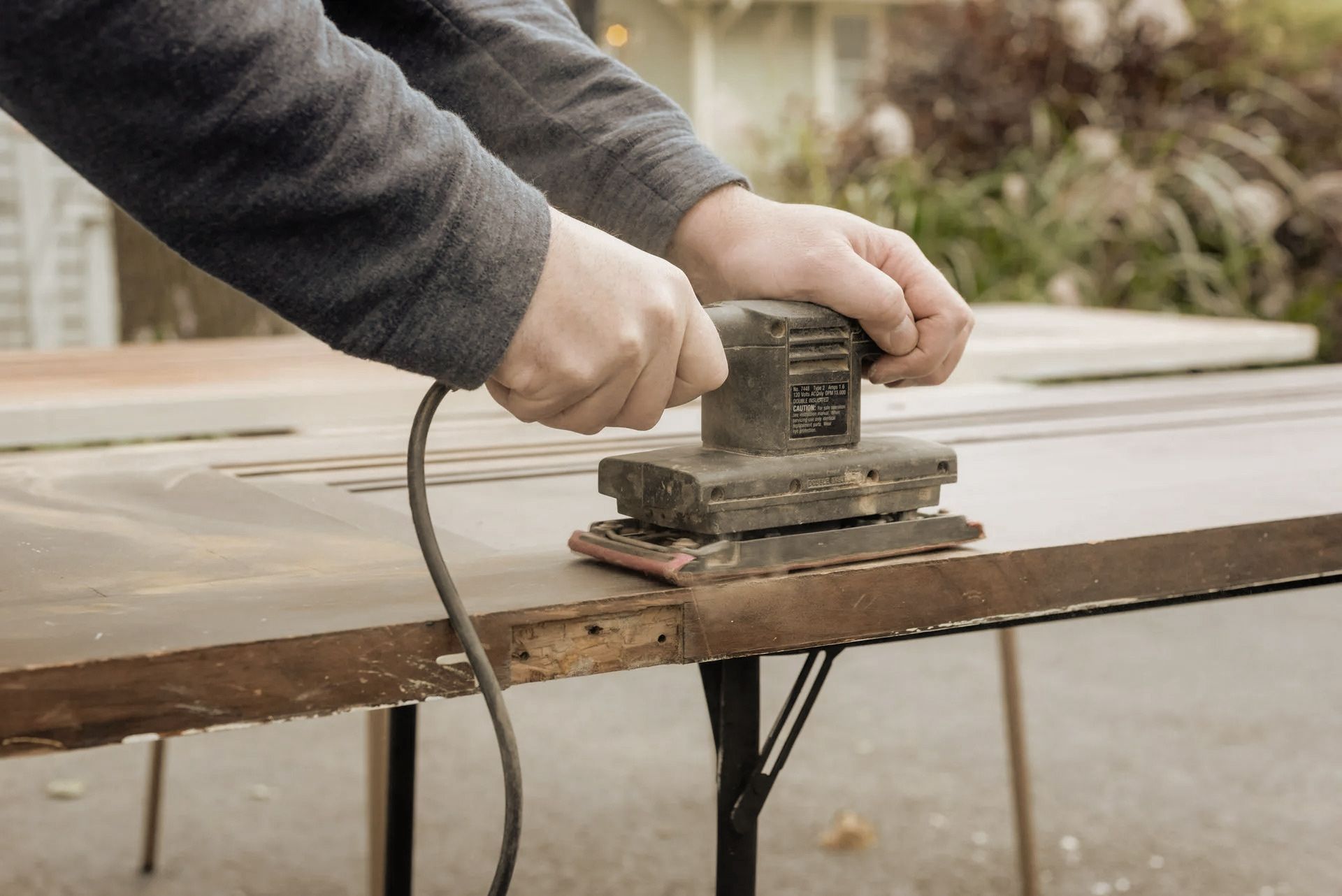 Person sands a wooden surface with an electric sander outdoors, likely a table.