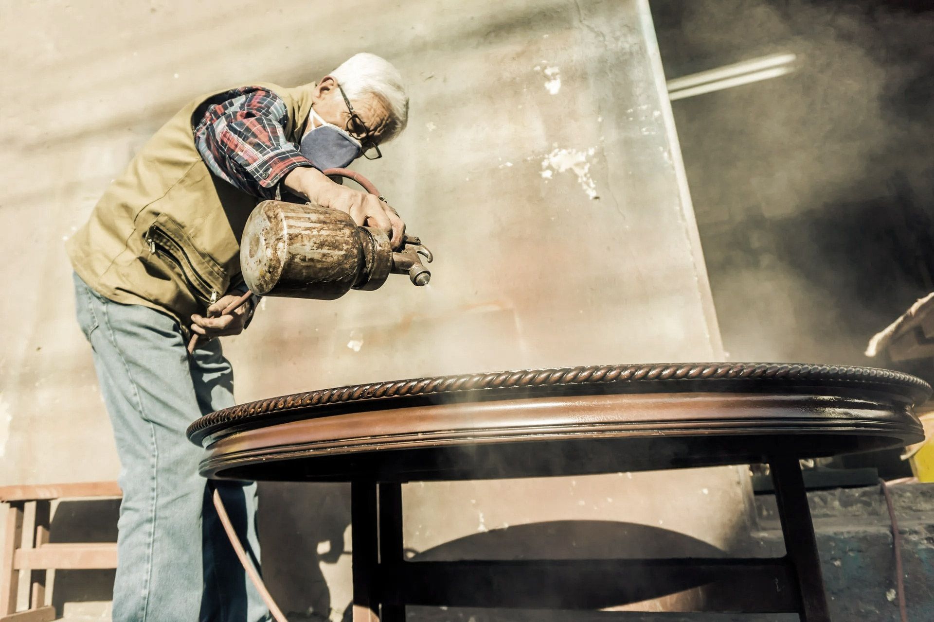 Man in mask sprays finish on a table in a workshop, generating a haze.