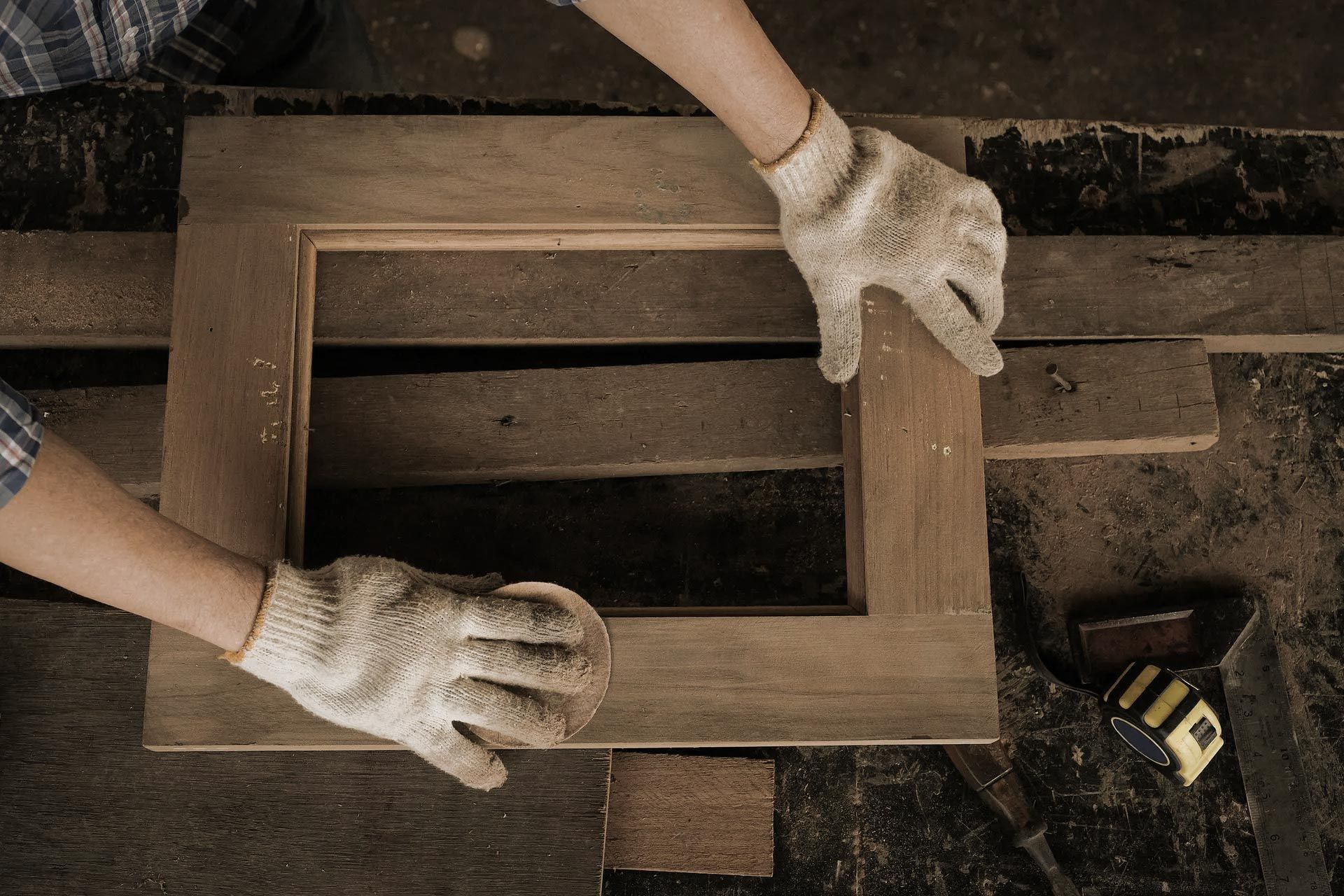 Hands wearing work gloves sanding a wooden frame on a workbench.