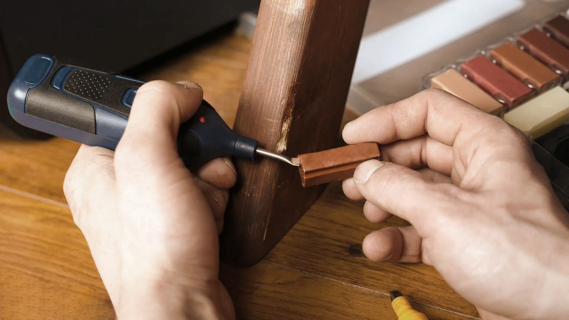 Person repairs furniture leg with a tool and a wood filler, on wooden surface.