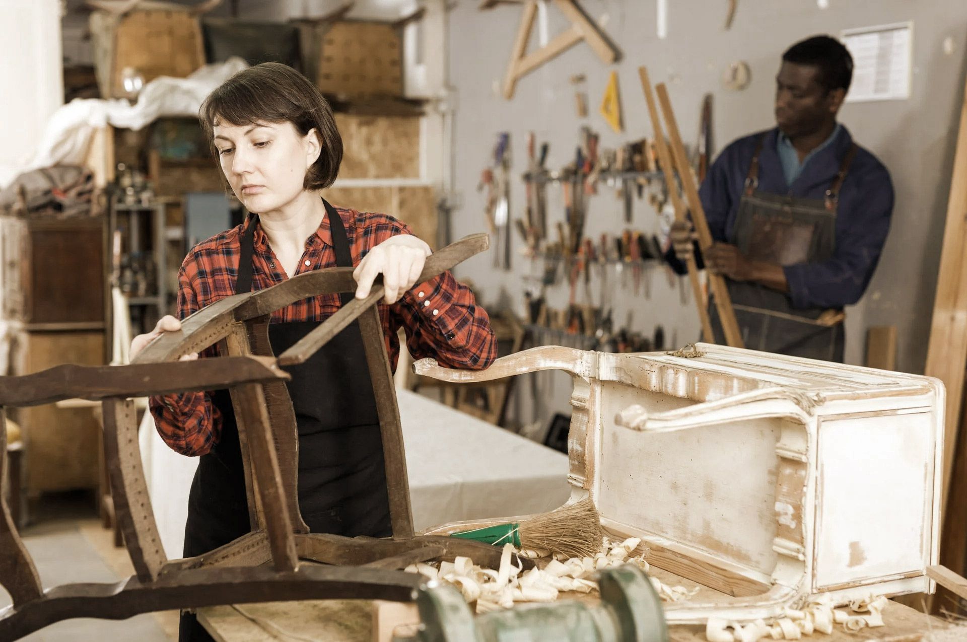 A person in workshop repairs furniture with another person watching.