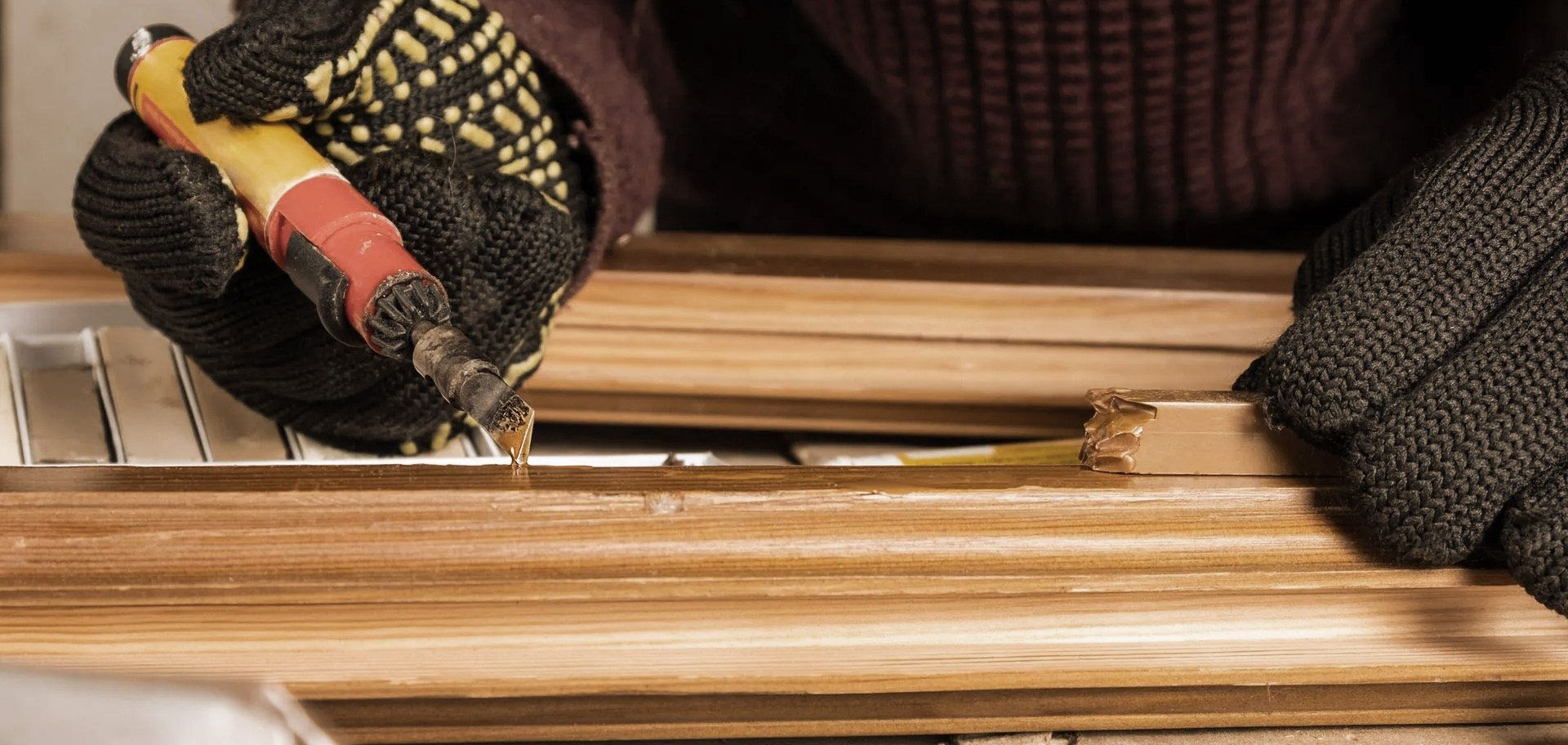 Person wearing gloves uses a tool to remove material from a wooden frame.