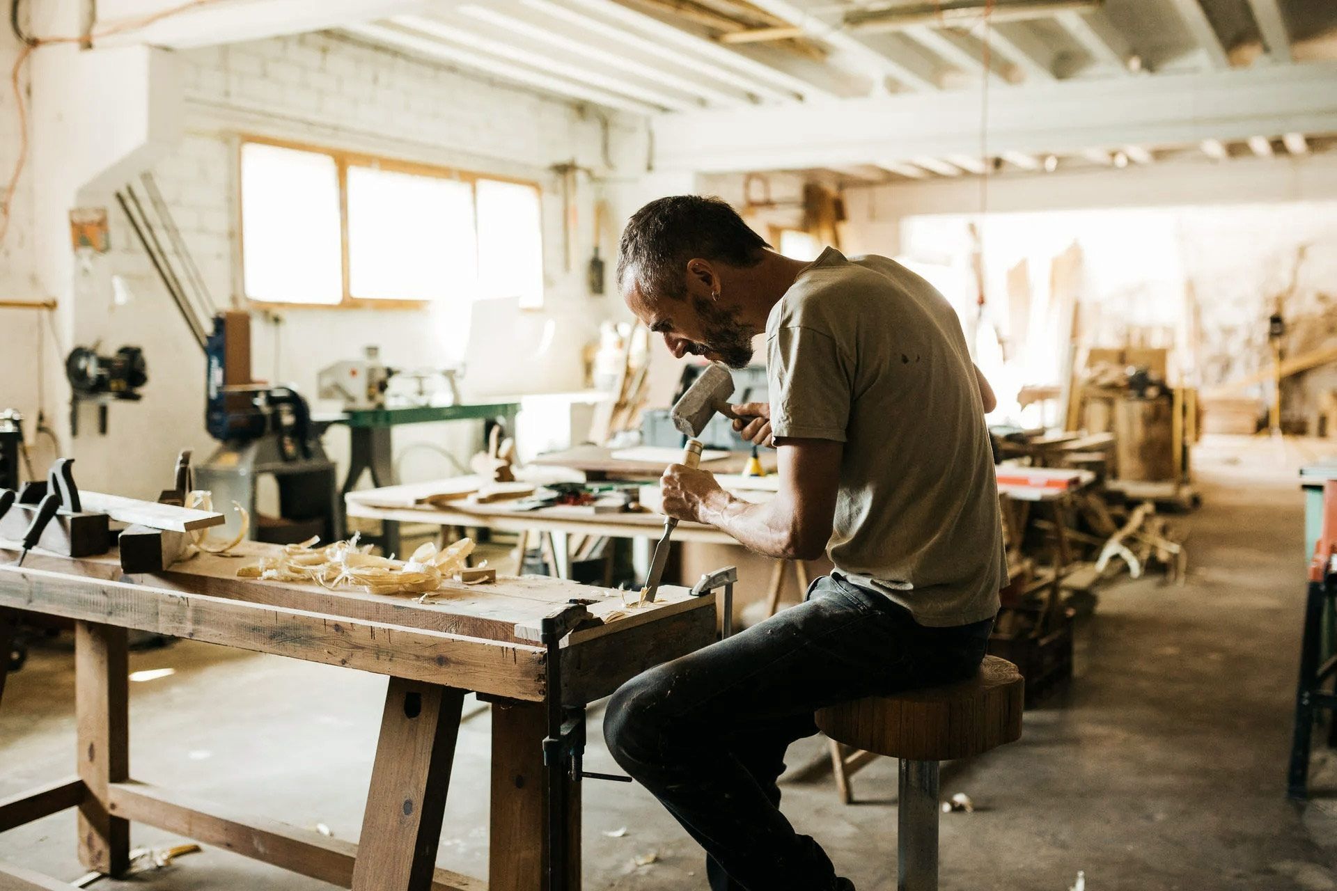 Woodworker carving wood at a workbench in a cluttered workshop.