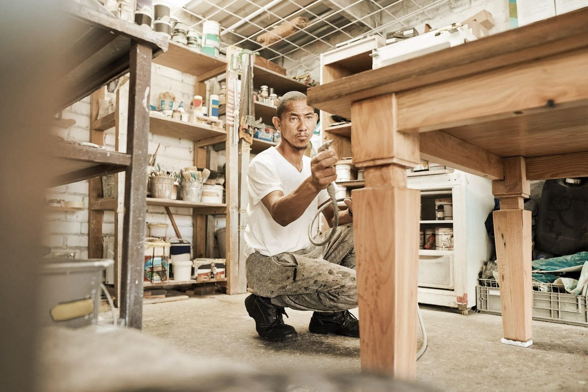 Man kneeling, working on wooden table leg in cluttered workshop.