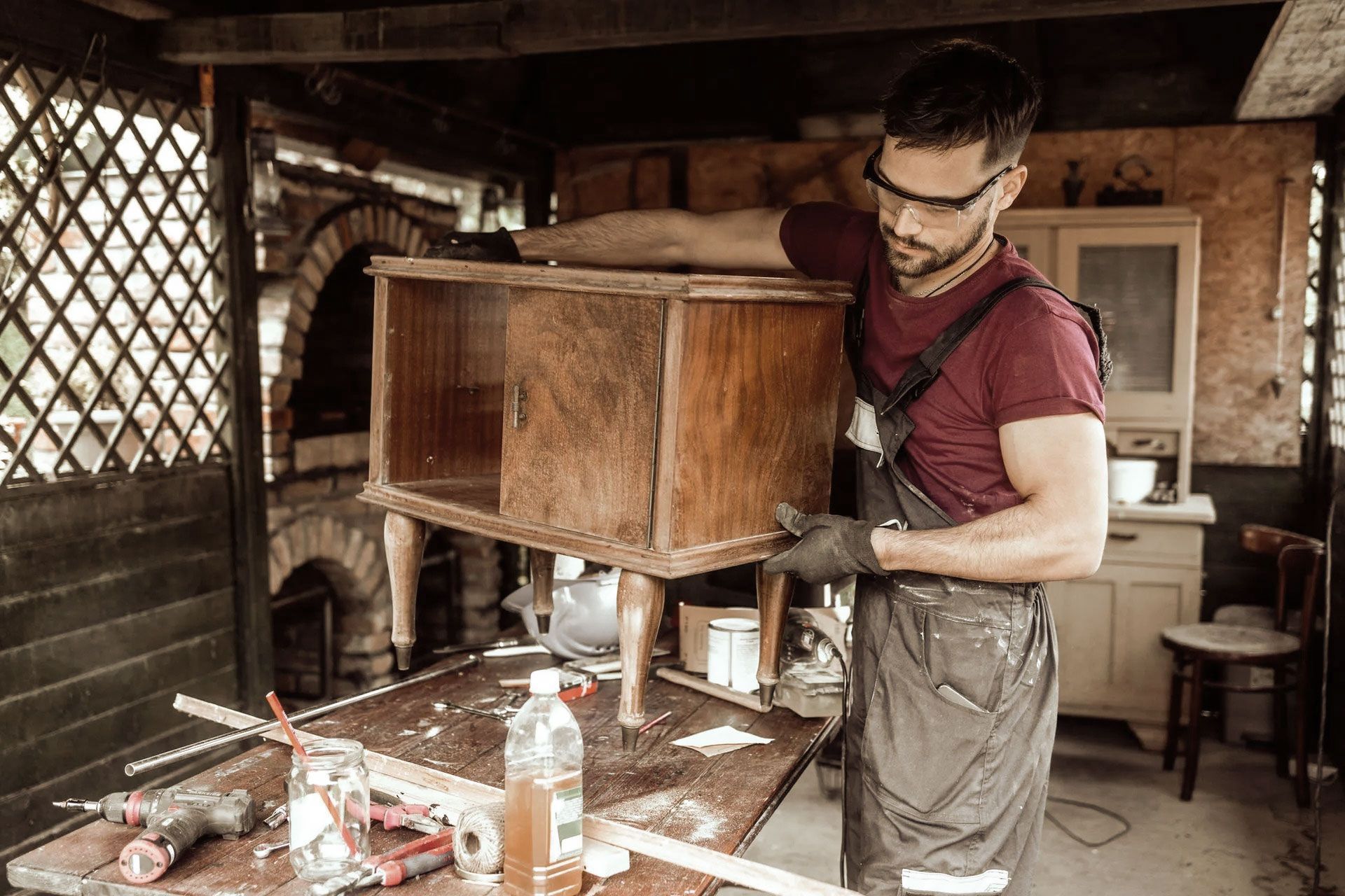 Man in overalls repairing wooden cabinet in a workshop; wearing safety glasses and gloves.