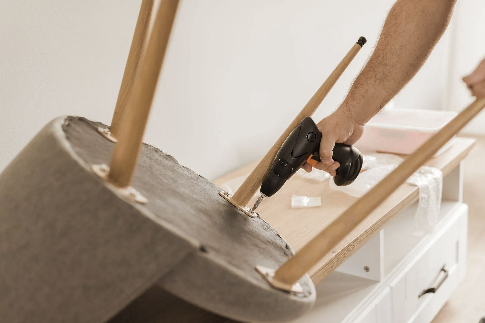Person using a power drill to attach a wooden leg to an overturned chair on a white cabinet.