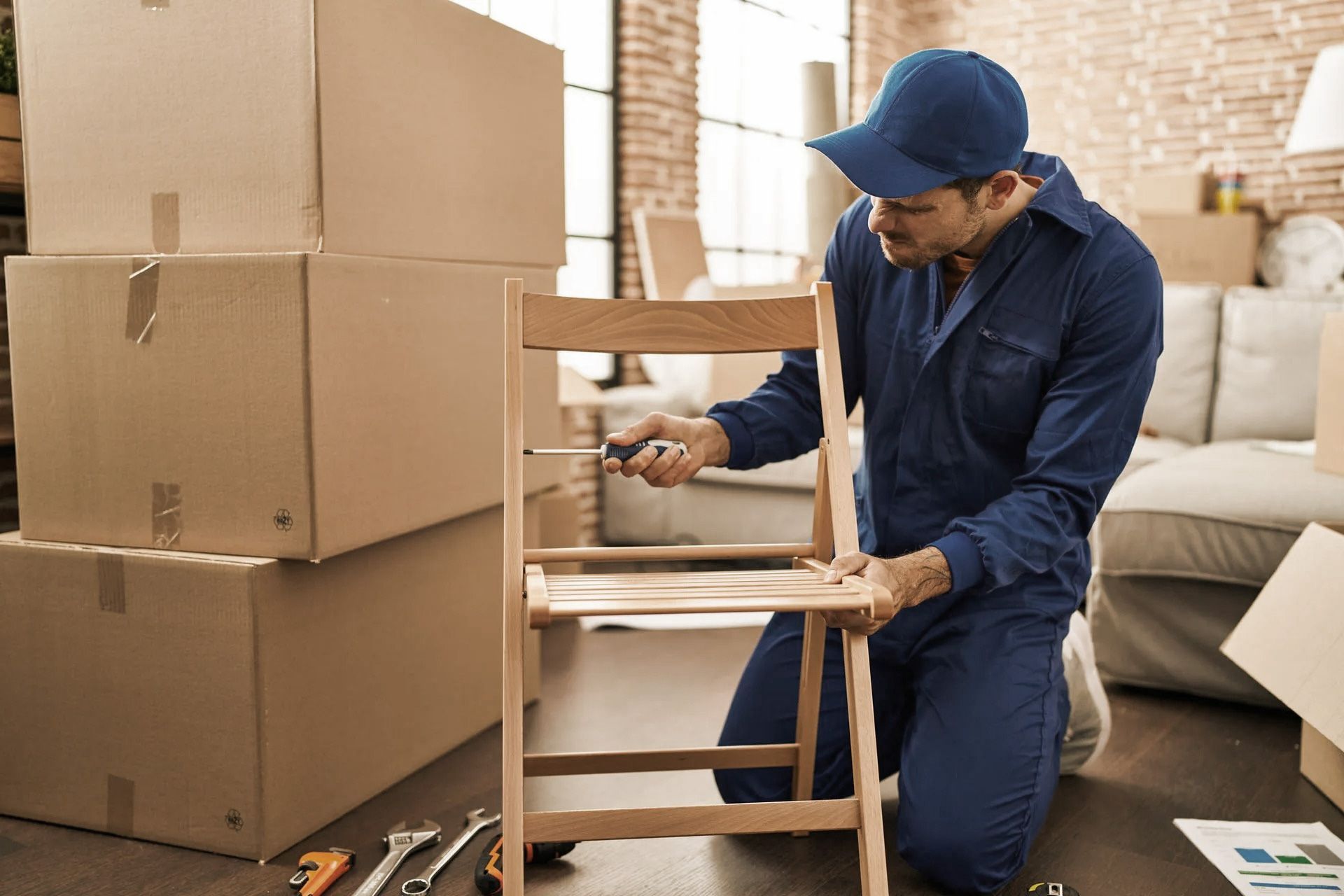 Person in blue jumpsuit assembles a chair with a screwdriver amid moving boxes in a room.