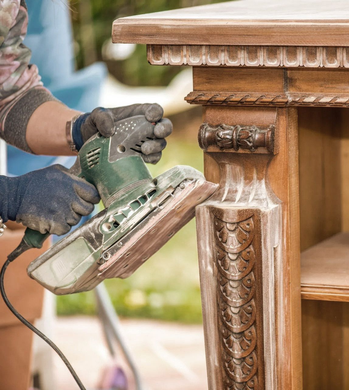 Person sanding an ornate wooden table with a power sander outdoors.