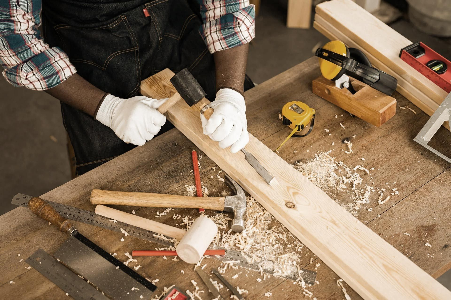 Person wearing gloves, carving wood with a chisel and mallet on a workbench.
