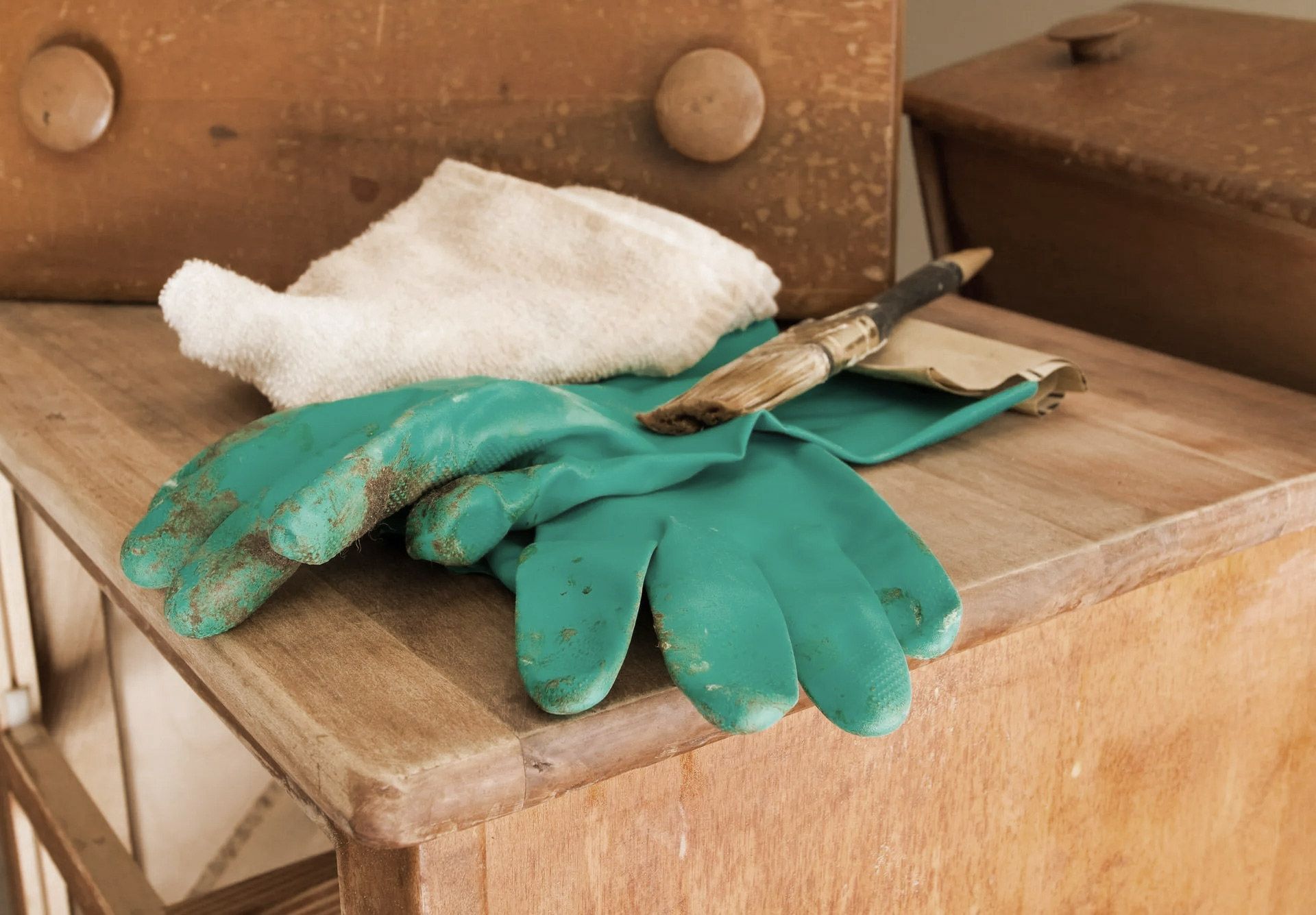 Green work gloves, rag, and paintbrush rest on a wooden surface with a drawer in the background.