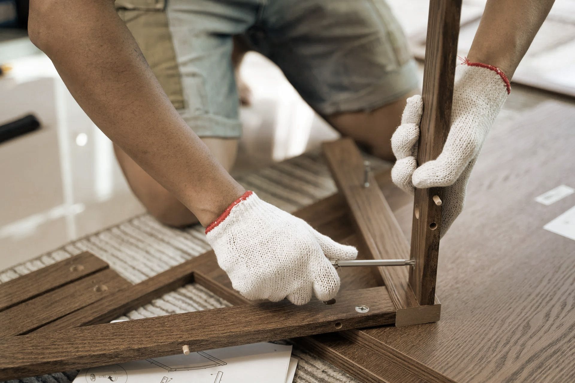Person assembling furniture, using a screwdriver, wearing gloves, indoors.