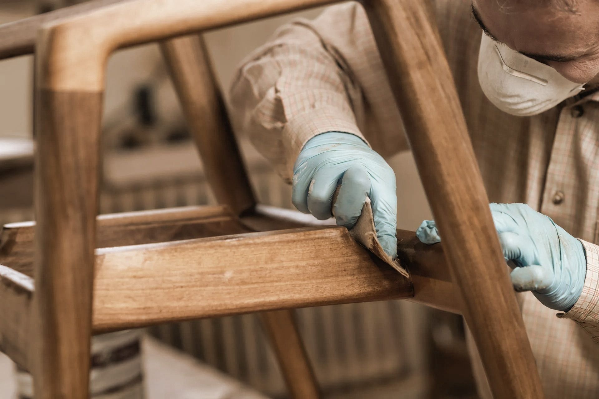 Person wearing a mask and gloves sanding a wooden chair indoors.