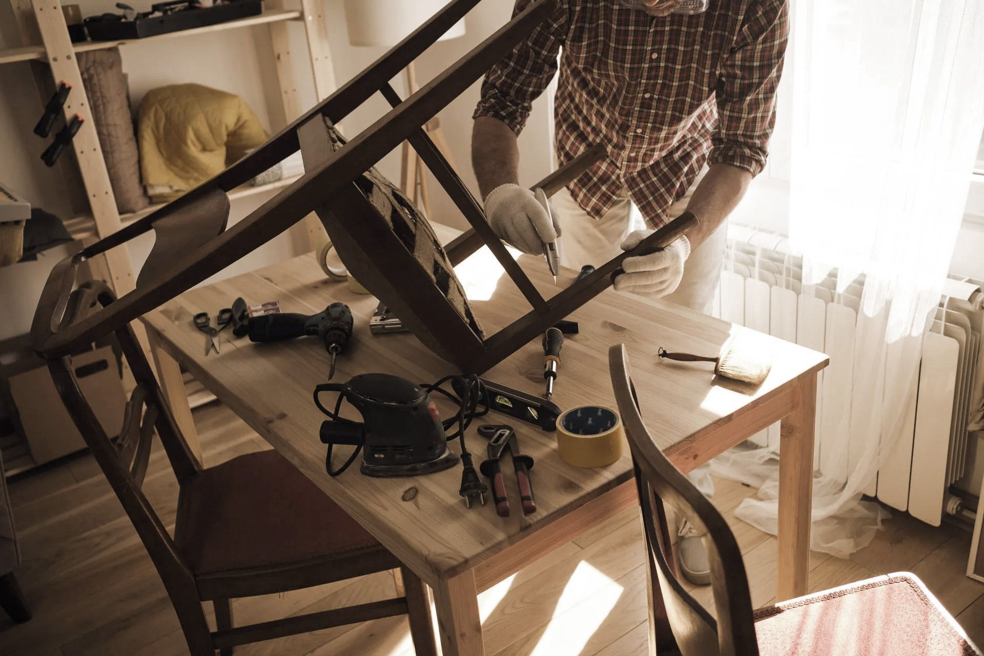 Person repairing a chair at a table with tools. Sunlight streams into the room.