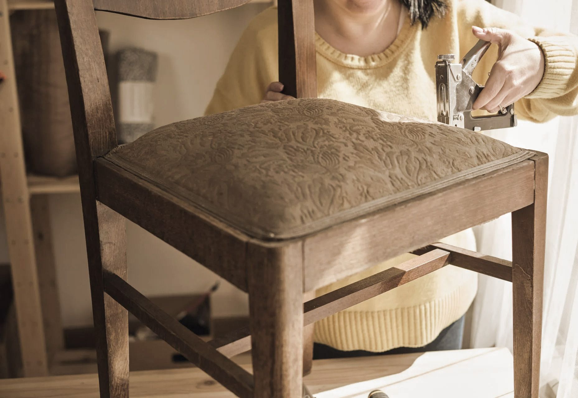 Person stapling fabric to a wooden chair seat indoors.