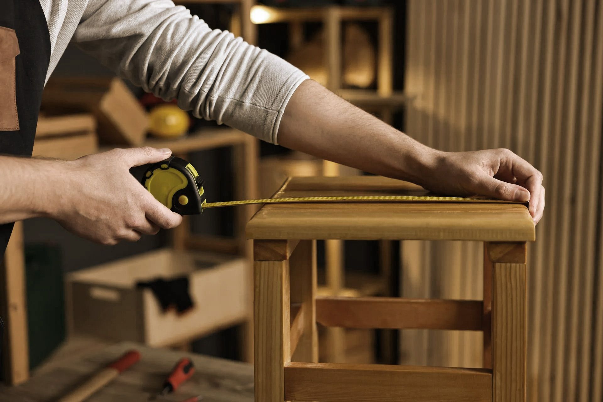 Person using a tape measure to measure the top of a wooden stool in a workshop.