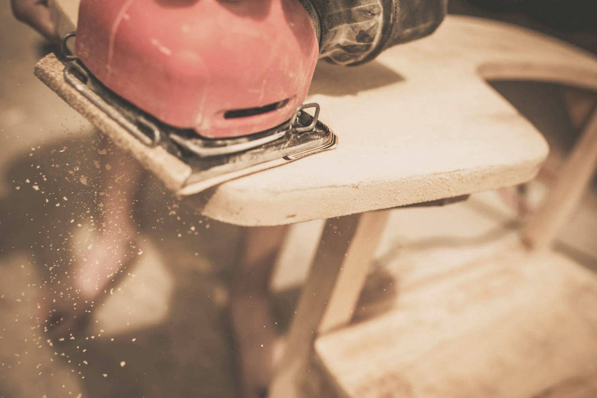 Sander smoothing wooden chair seat; sawdust visible.