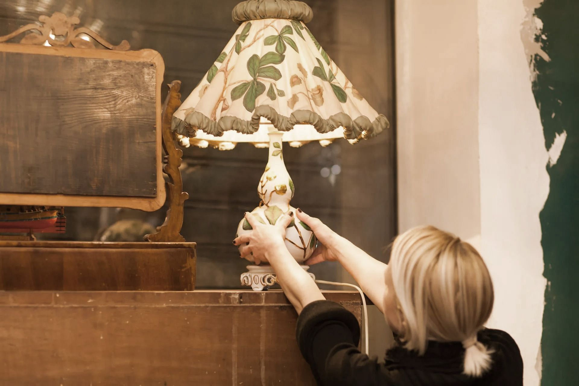 Woman adjusting a lamp on a dresser with a mirror, interior setting.