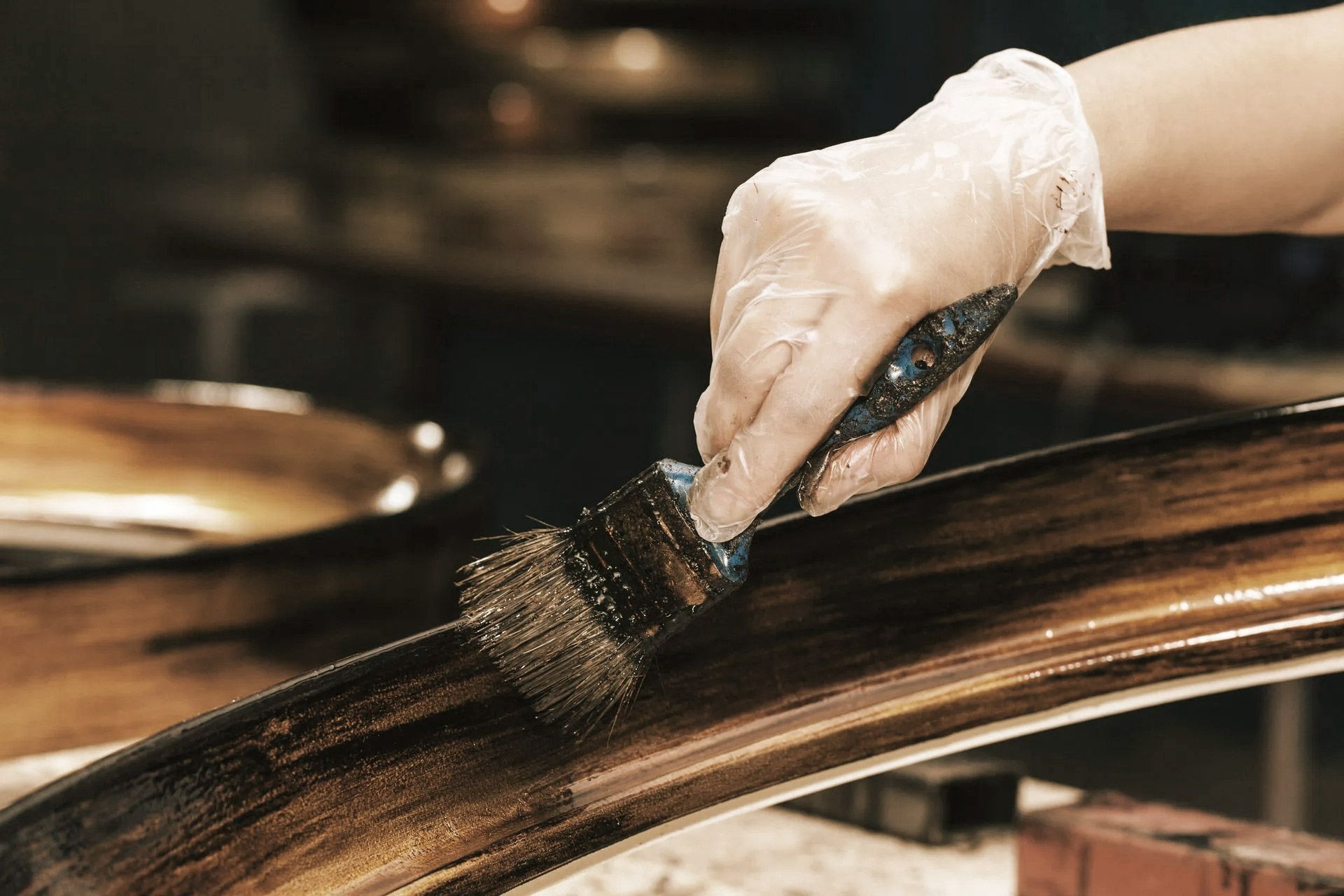 A gloved hand uses a brush to apply dark paint to a curved wooden frame.