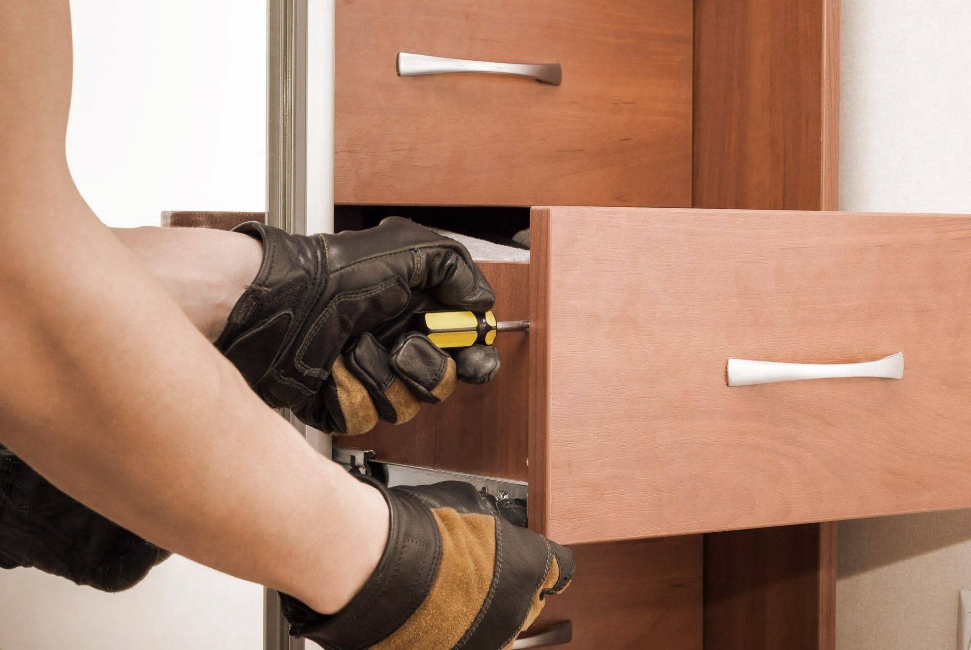 Person wearing gloves using a screwdriver to adjust a drawer in a wooden cabinet.