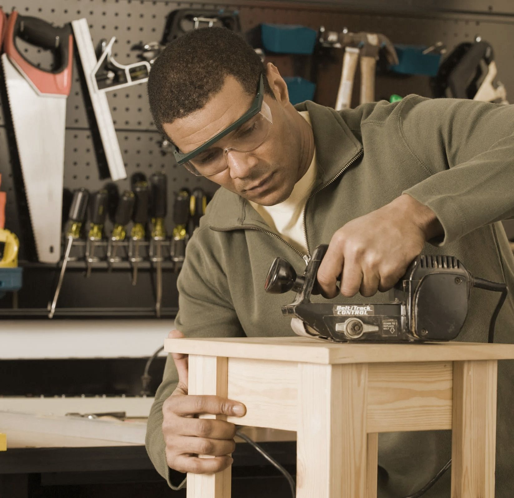 Man using a power sander on a wooden stool in a workshop, wearing safety glasses.