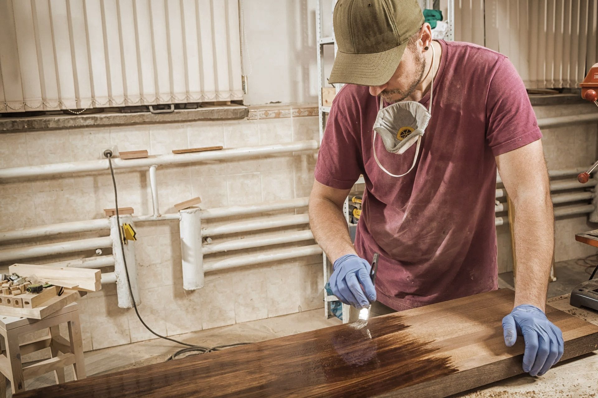 Man wearing gloves and respirator applying finish to wood in a workshop.