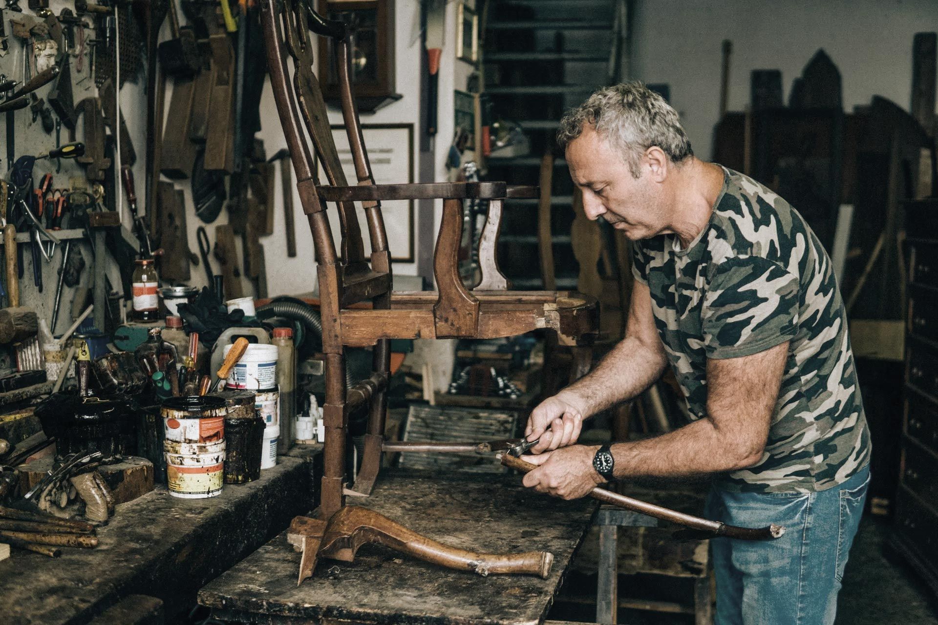 Man repairs antique wooden chair in a cluttered workshop, wearing camouflage shirt.