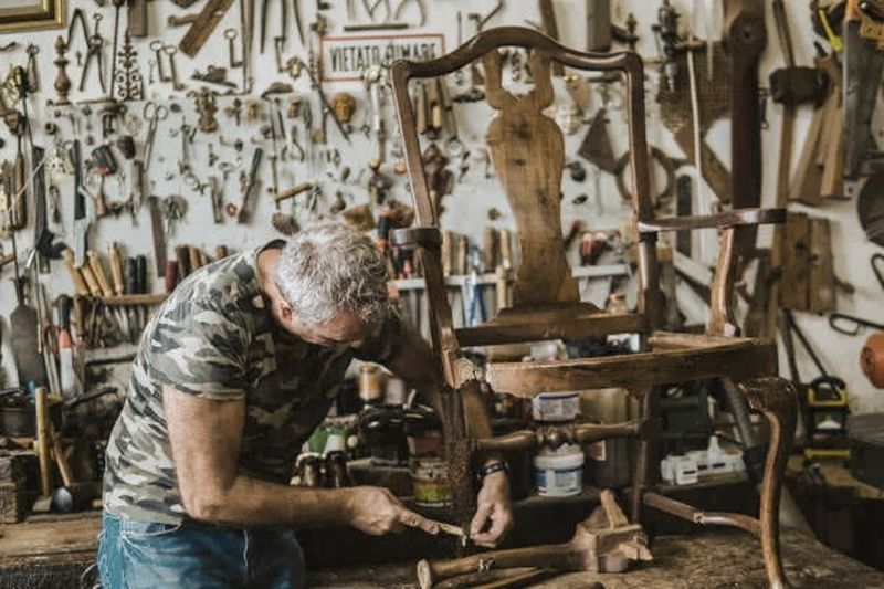 Man working on a wooden chair in a workshop with tools hanging on the wall.