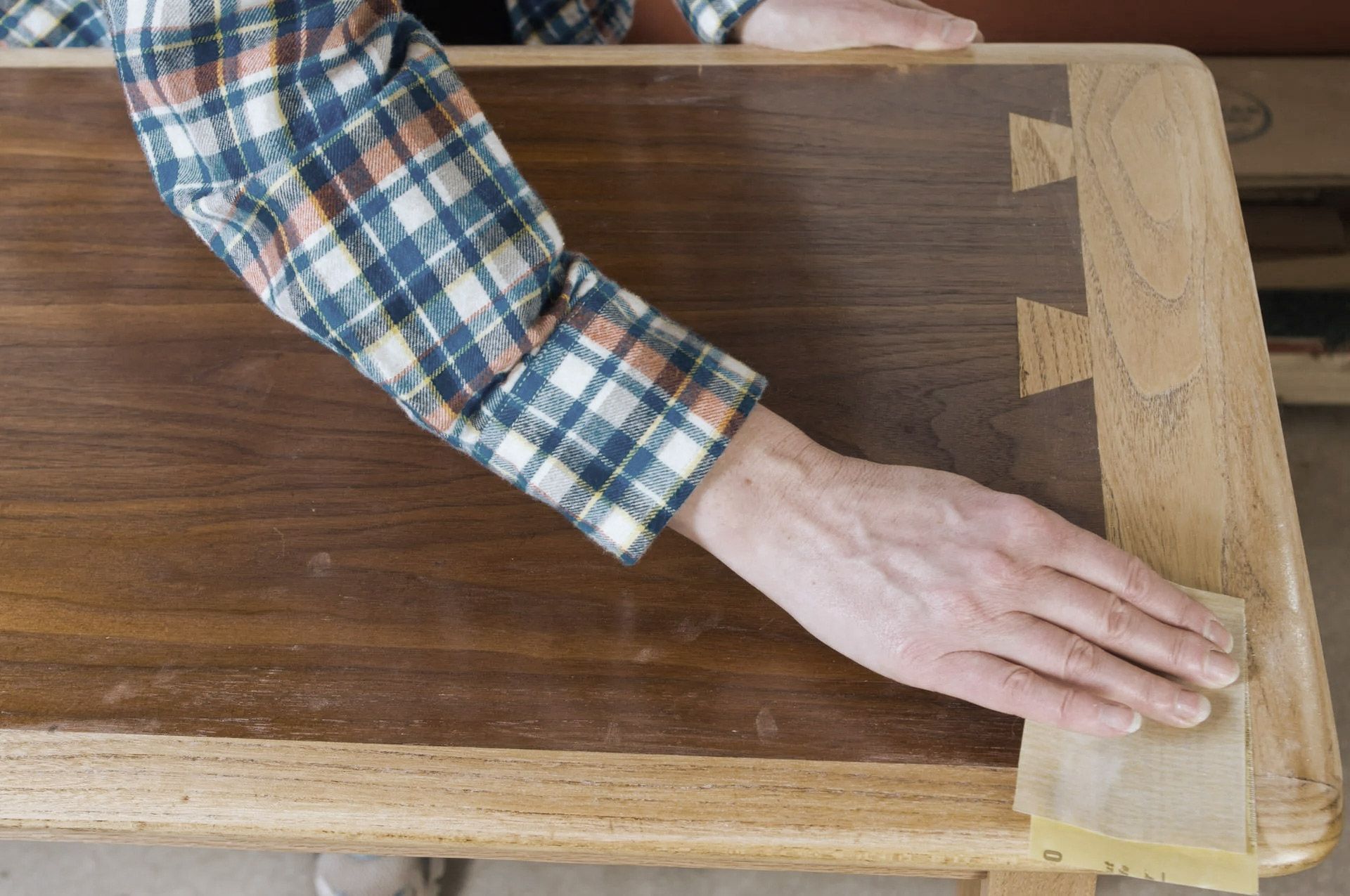 Person sanding a wooden table with exposed dovetail joints, wearing a plaid shirt.