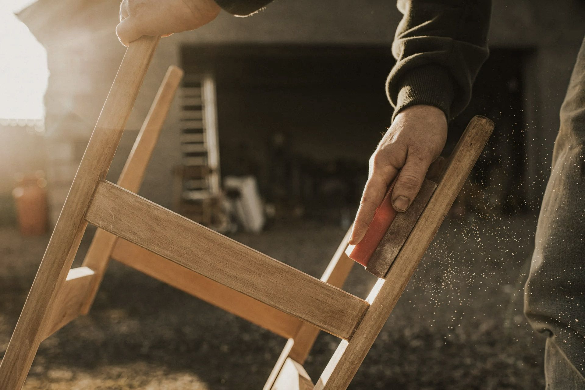 Person sanding a wooden chair outdoors; sunlight, visible workshop tools in background.