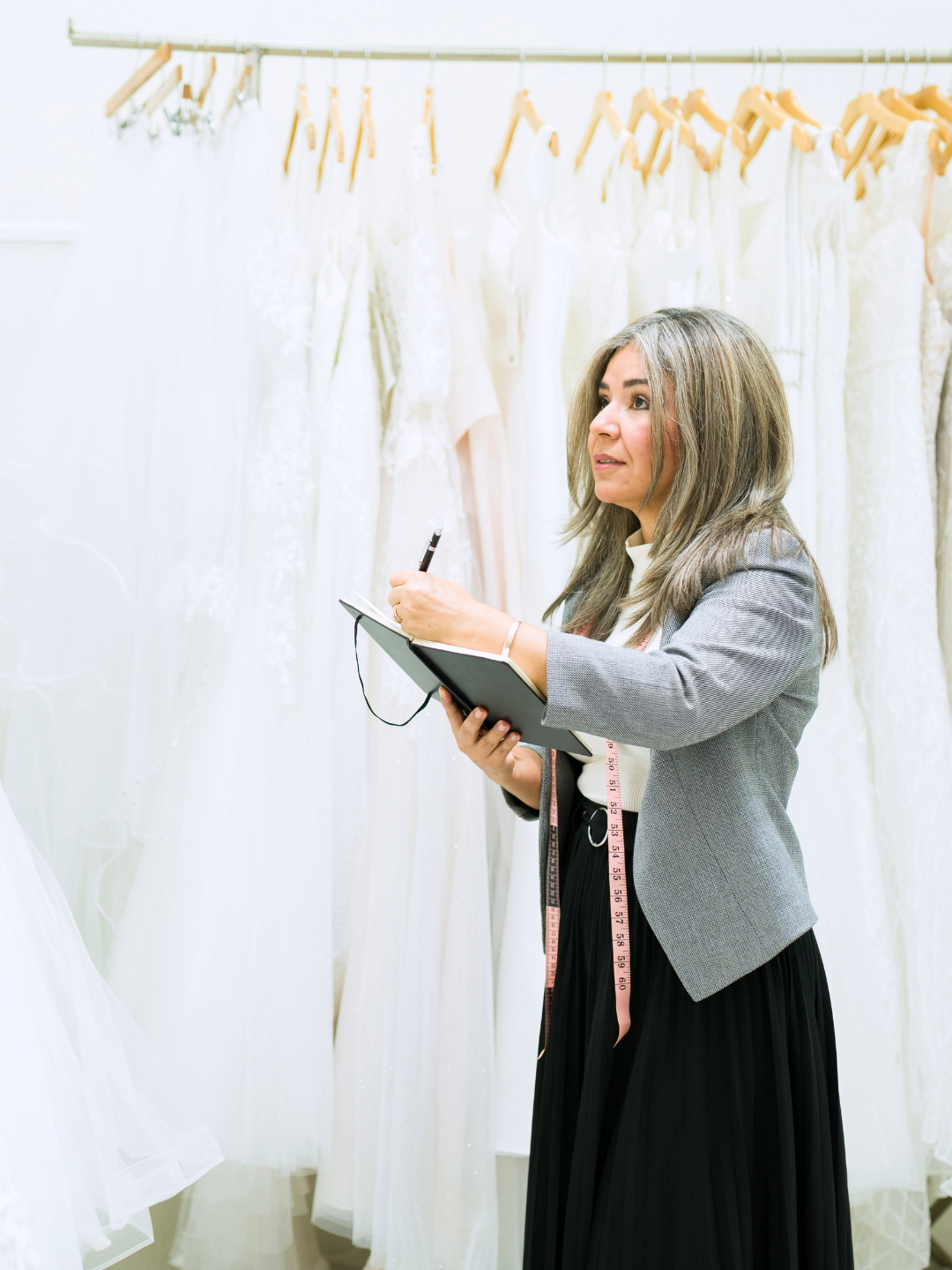 Woman in a blazer taking notes while looking at a row of white wedding dresses.