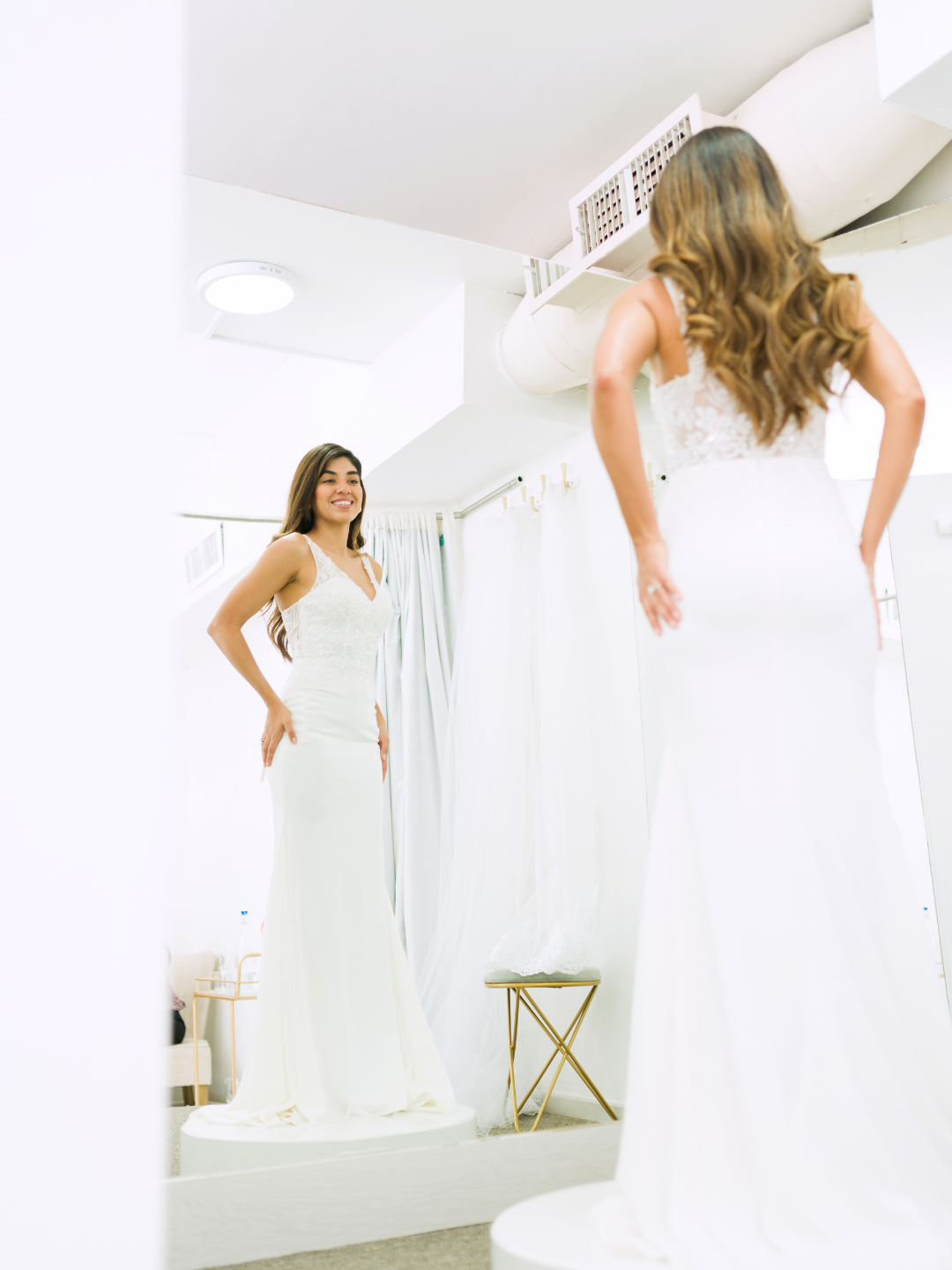 Woman in wedding dress looking in mirror, smiling. Bridal shop setting.