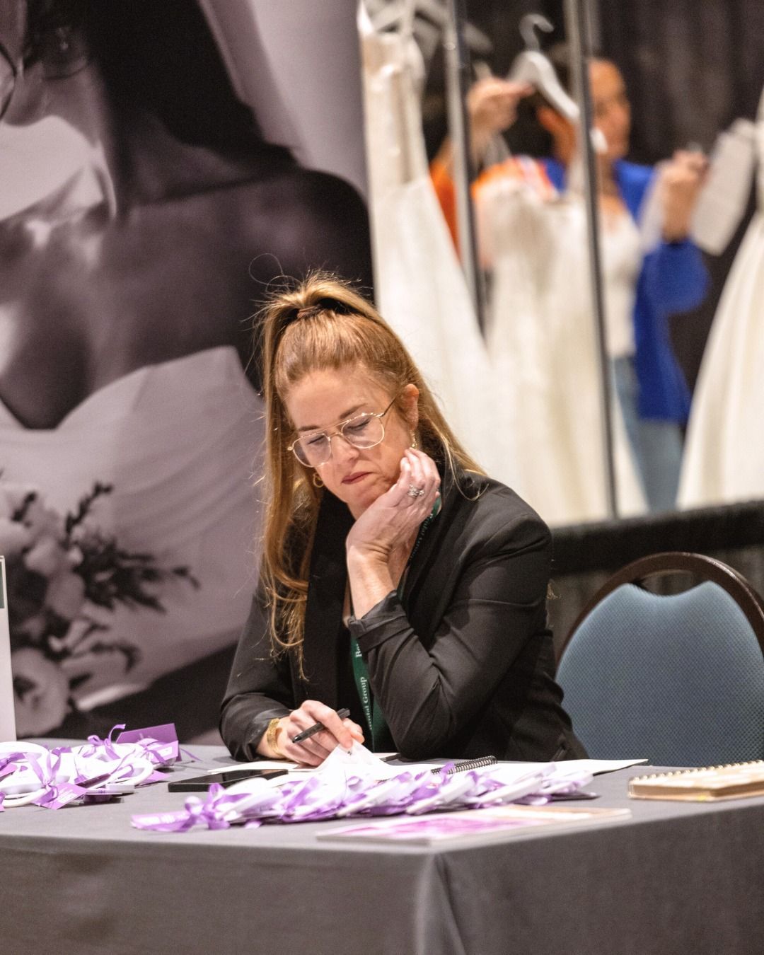 Woman with glasses at table, looking at papers. Wedding dress display in background.