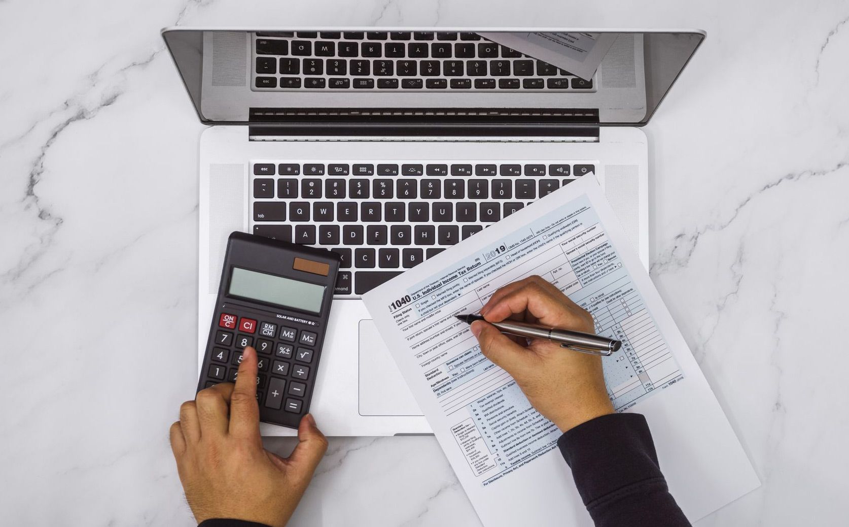 A person is using a calculator and writing on a piece of paper in front of a laptop computer.