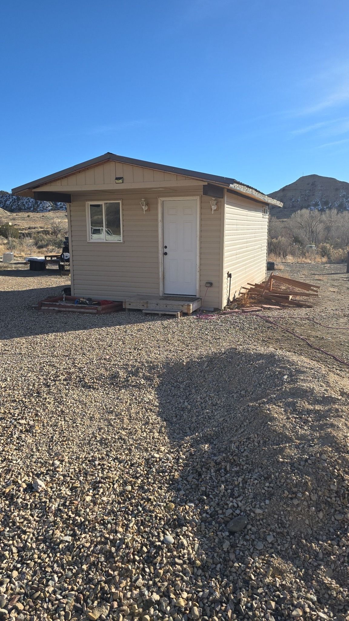 Small beige shed with a white door and window sits on gravel in a desert landscape.
