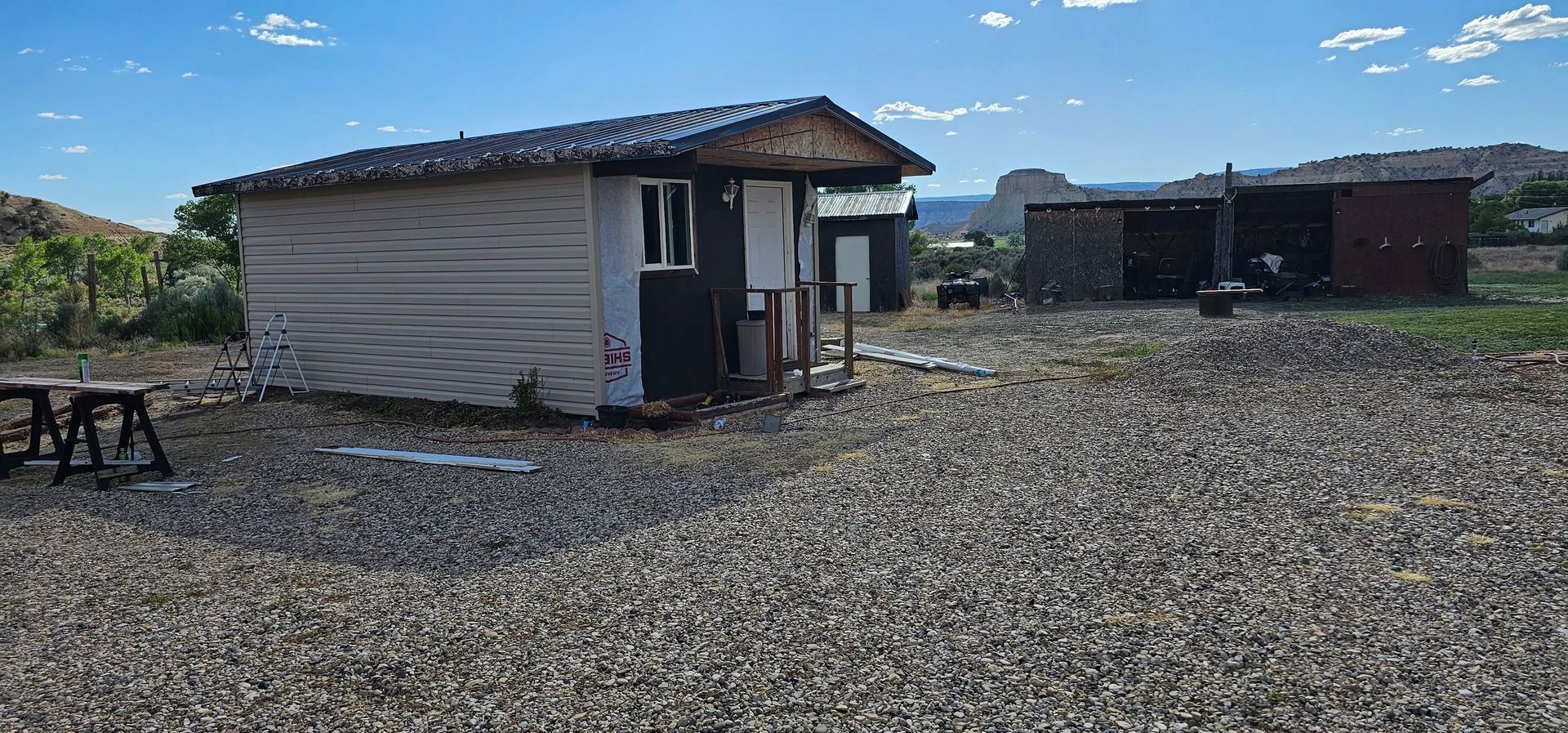 A small, weathered house and outbuildings sit on a gravel lot under a blue sky, with distant hills.