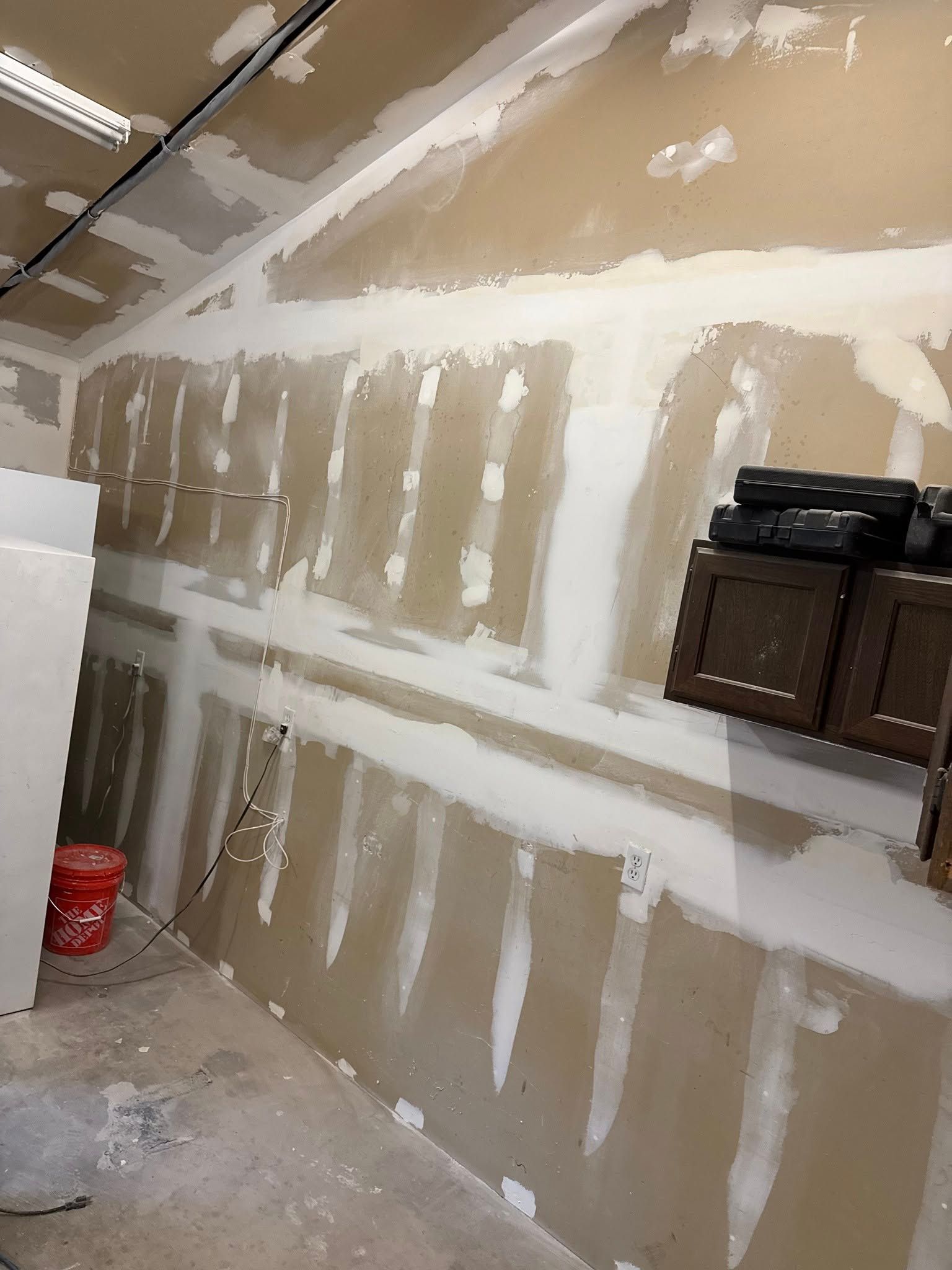 Interior wall with drywall installed, mudded and partially finished; cabinets and a bucket present.