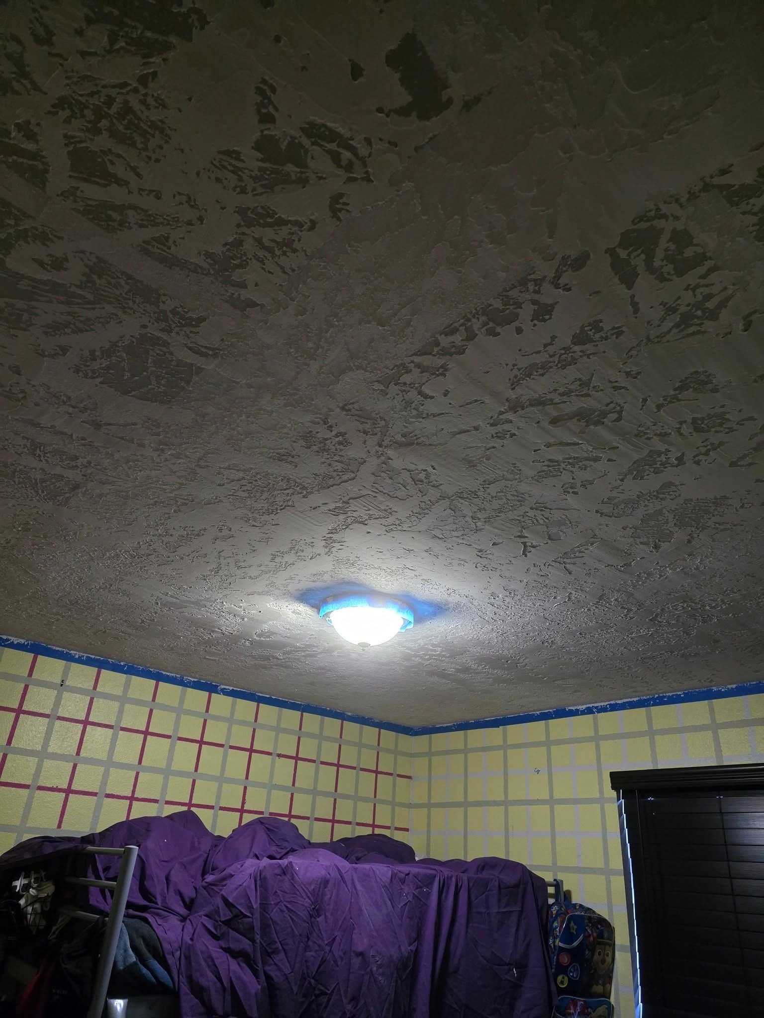 Room ceiling with popcorn texture, a central light, and taped blue border. Grid-lined yellow wall. Purple fabric and black cabinet.