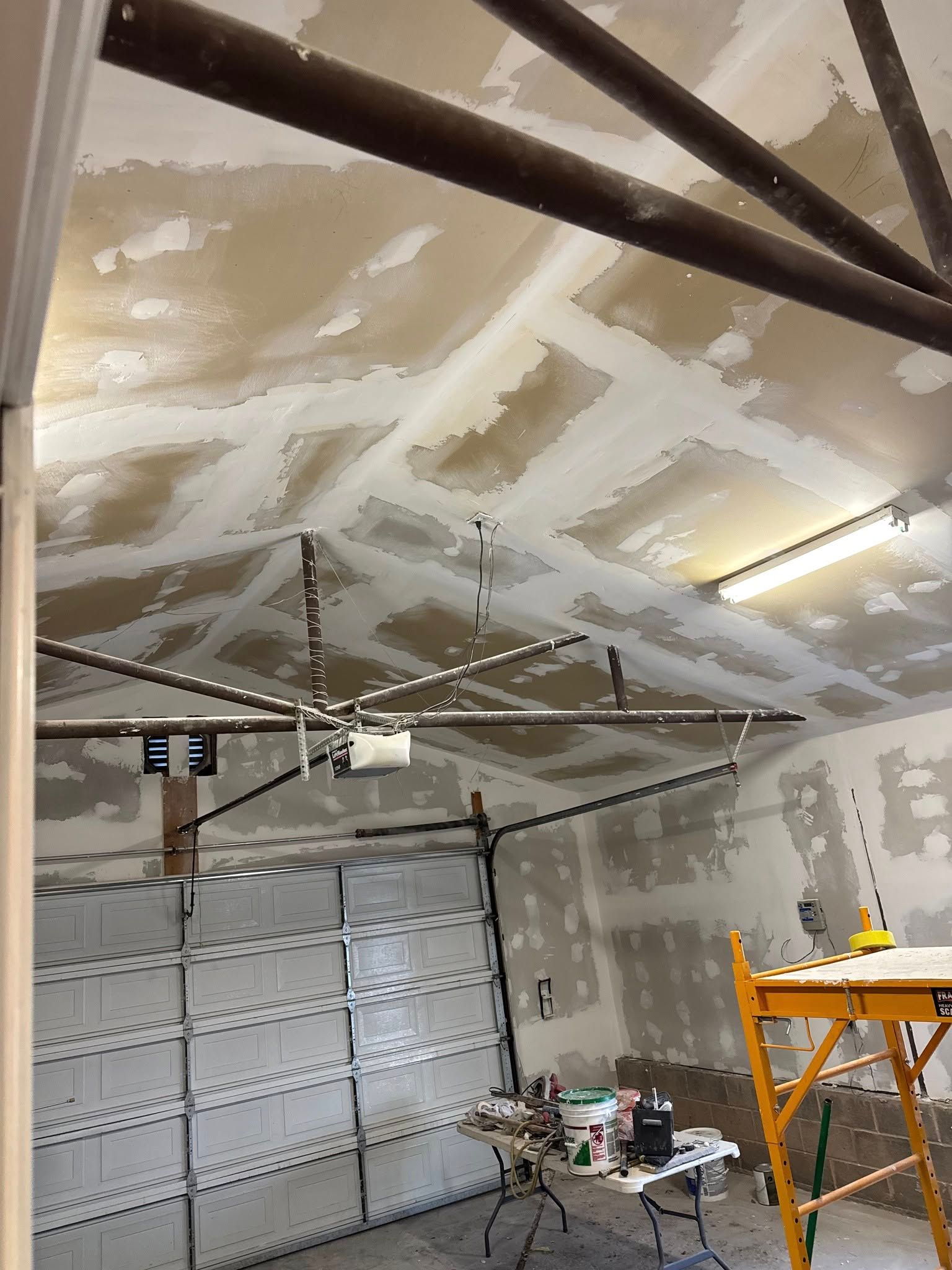 Garage ceiling with drywall mud applied, partially covering the textured surface. Brown beams and garage door visible.