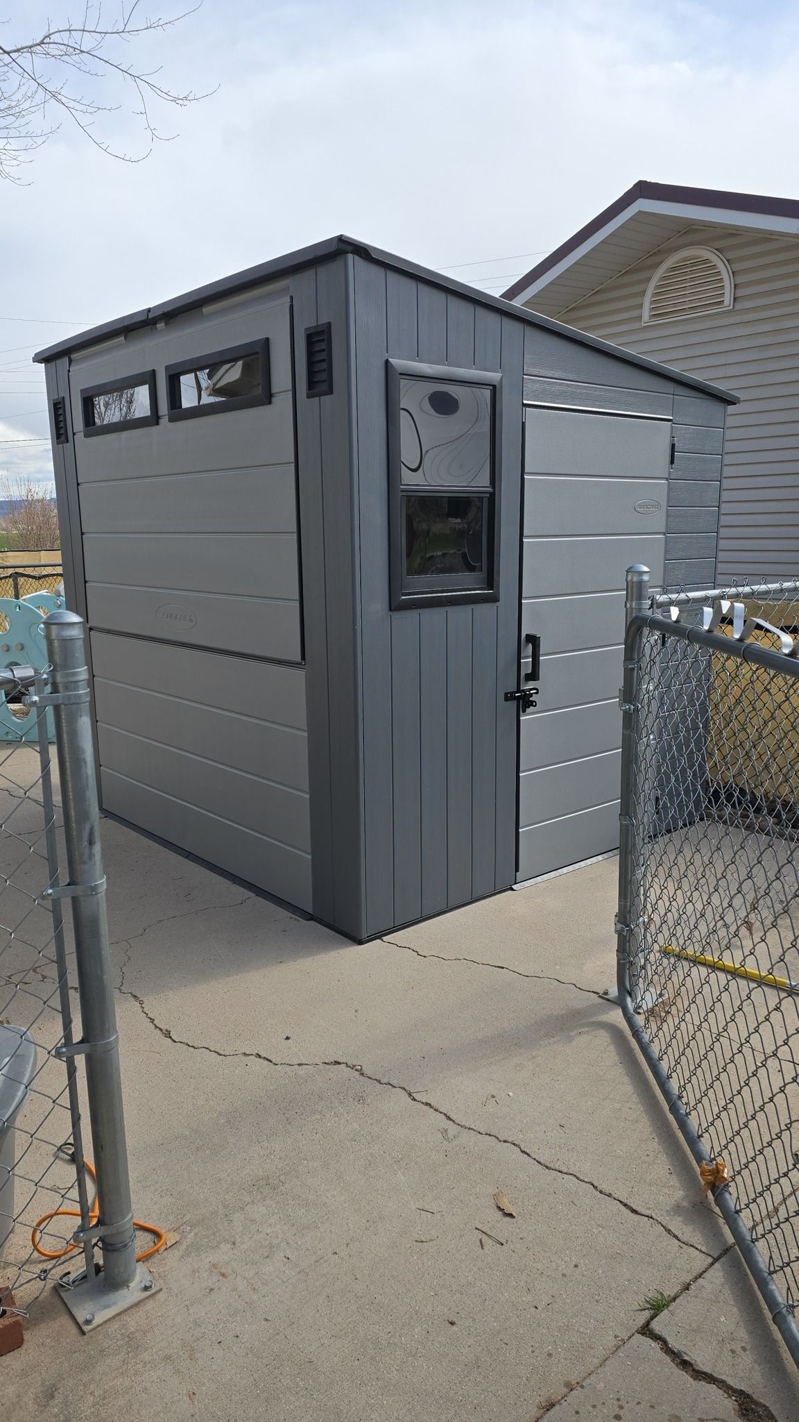 Gray shed with dark trim, windows, and a door, next to a chain-link fence and a building.