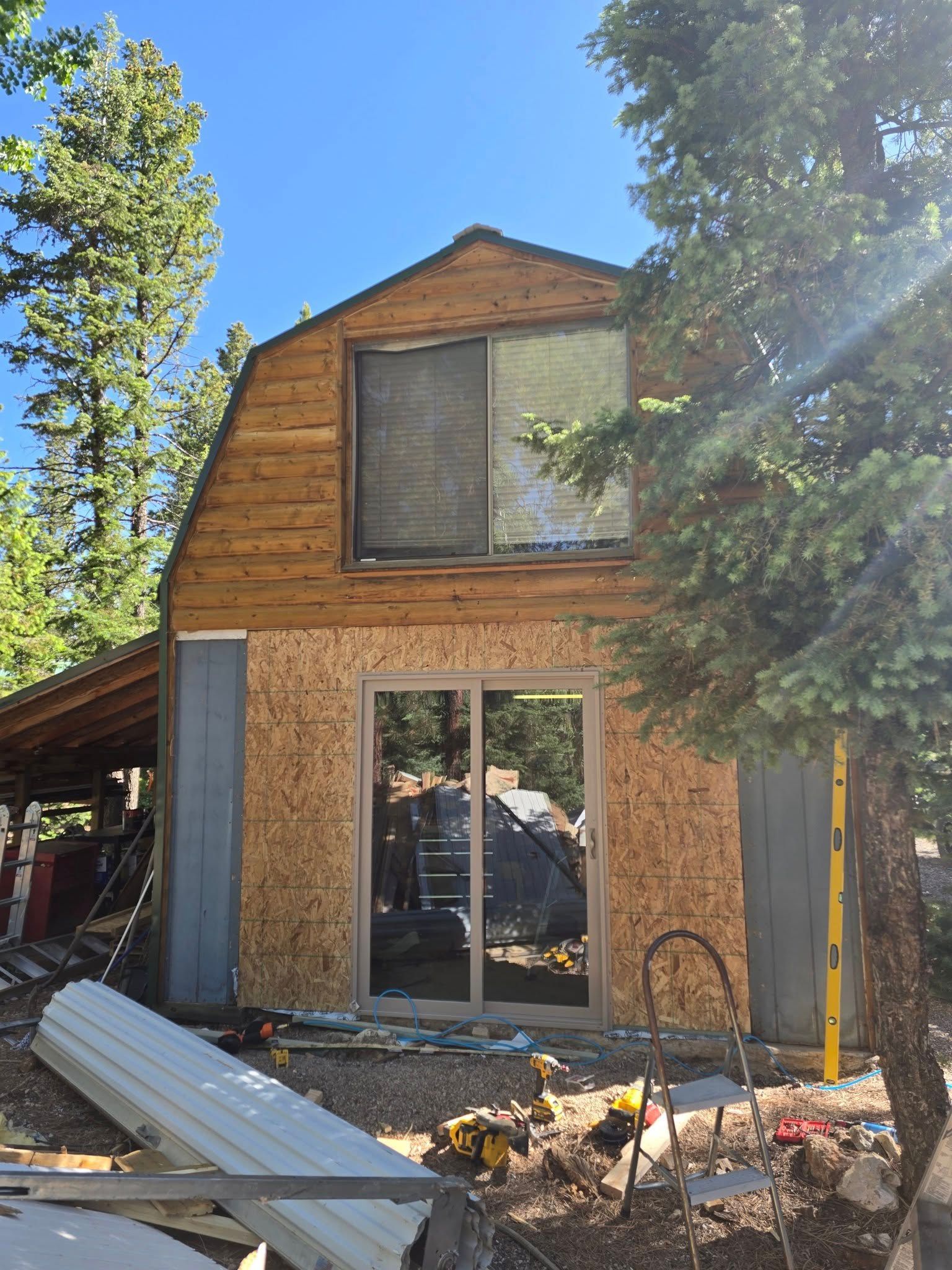 Barn-shaped building under construction with exposed wood, windows, and surrounding trees on a sunny day.
