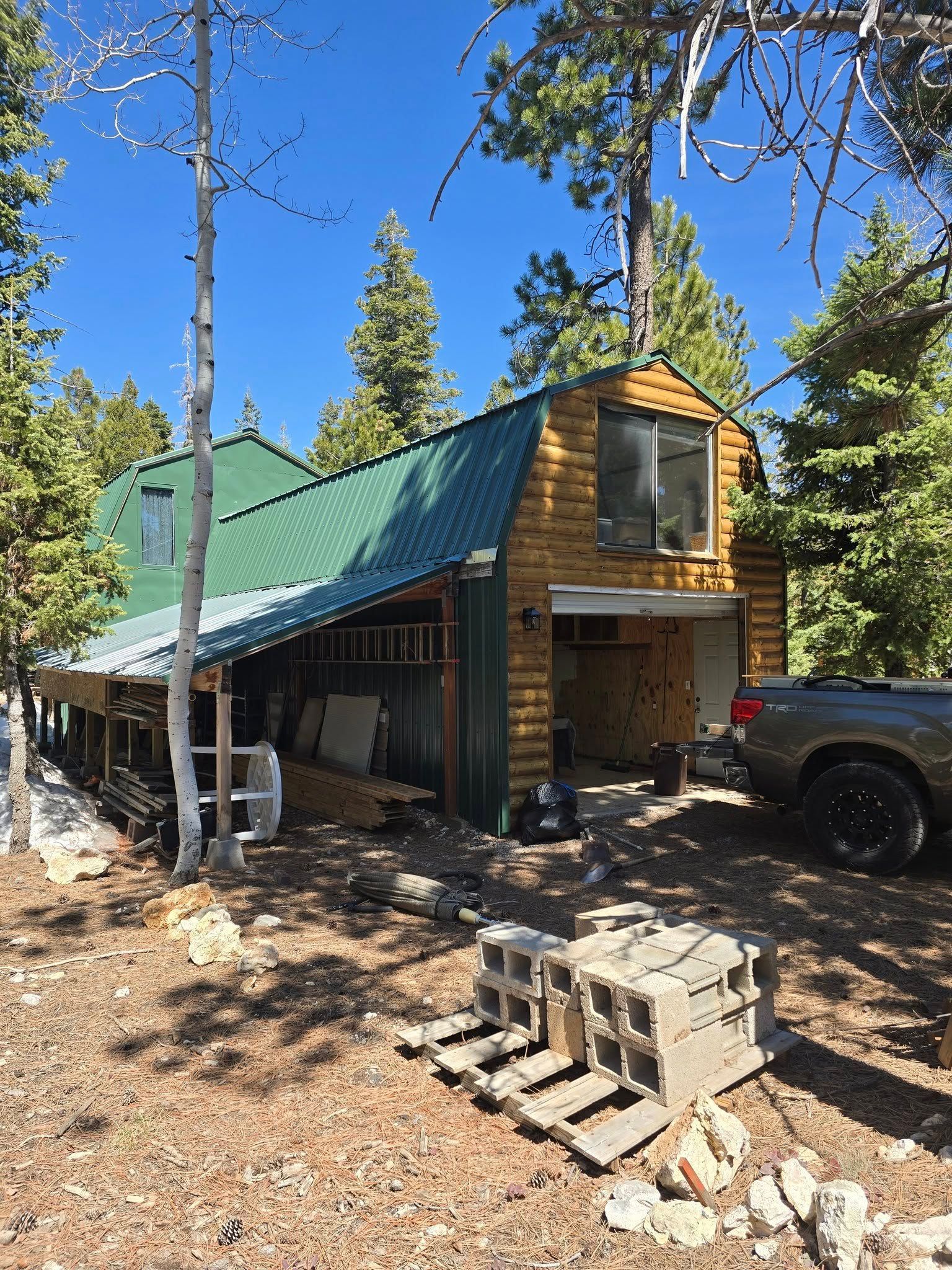 A wooden garage with green roof, a pickup truck in the driveway, surrounded by trees, and construction materials.
