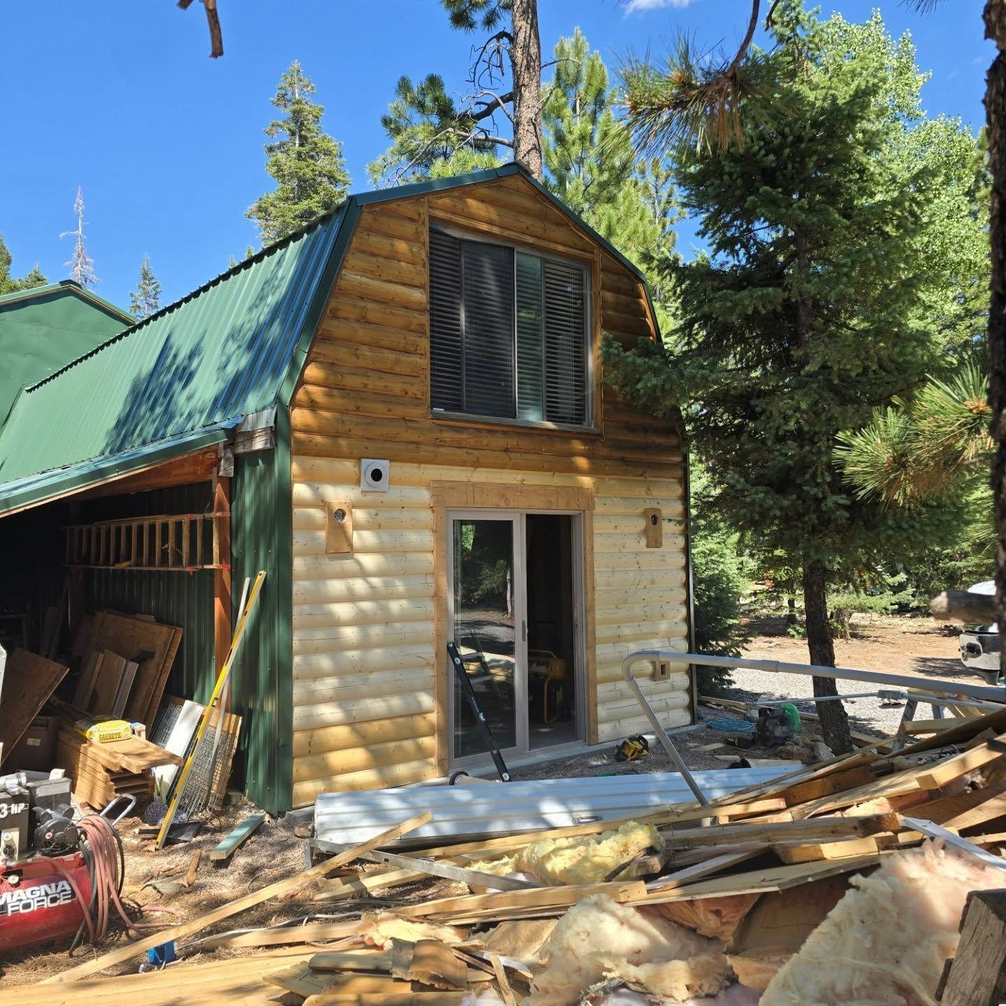 A cabin with green roof and light wood siding, under construction surrounded by debris, in a forest setting.