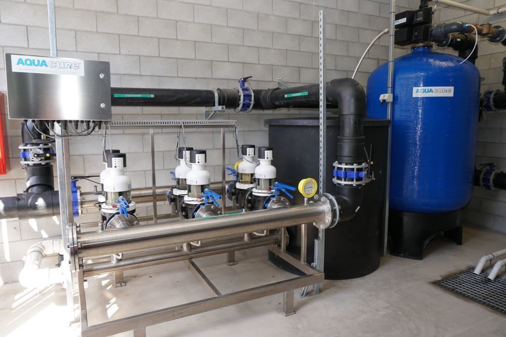 Water filtration system with blue tank, black pipes, and stainless steel components against a gray brick wall.