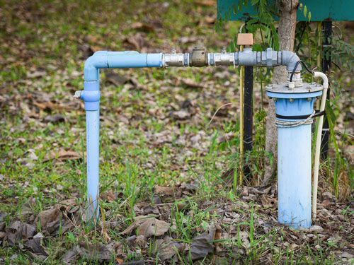Blue water well pipe system in a grassy yard, connected by metal and plastic fittings.