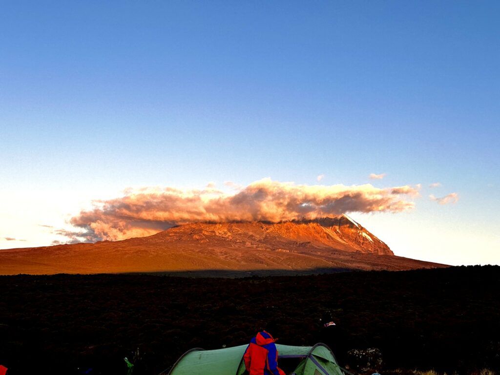 A person is standing in front of a tent with a mountain in the background.