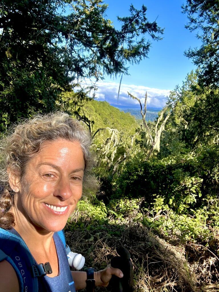A woman is smiling while hiking in the woods.